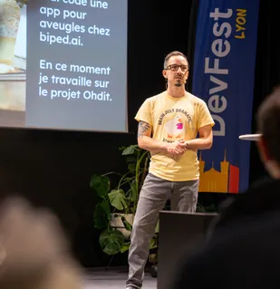 Photo de Gabriel Pillet en train de présenter une conférence sur l'accessibilité numérique au DevFest Lyon 2025. Sur son t-shirt figure un canard qui tiens une tasse de café, avec un texte anglais qu'on peut traduire par "Cerveau parfois un peu dramatique".